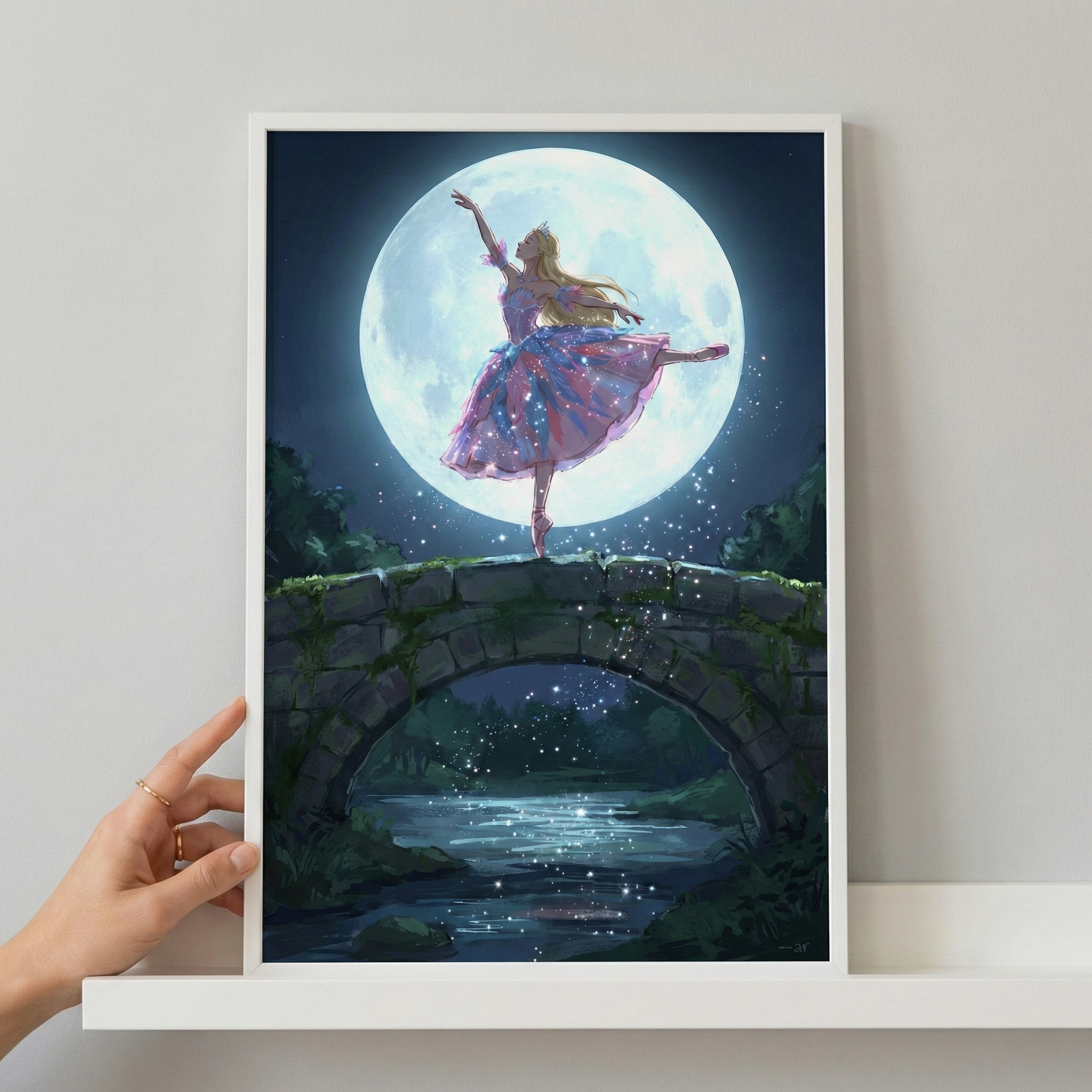 A graceful female figure in a ballet pose stands on one foot atop a mossy stone bridge. She wears a shimmering, multi-layered dress in shades of pink, purple, and blue, adorned with glowing sparkles. Her arms are raised elegantly as she dances against the backdrop of an enormous, luminous full moon that fills the dark night sky. Below the bridge, a calm river reflects the moonlight and a trail of glowing particles. The artwork is in a white frame, held by a hand on a white shelf.