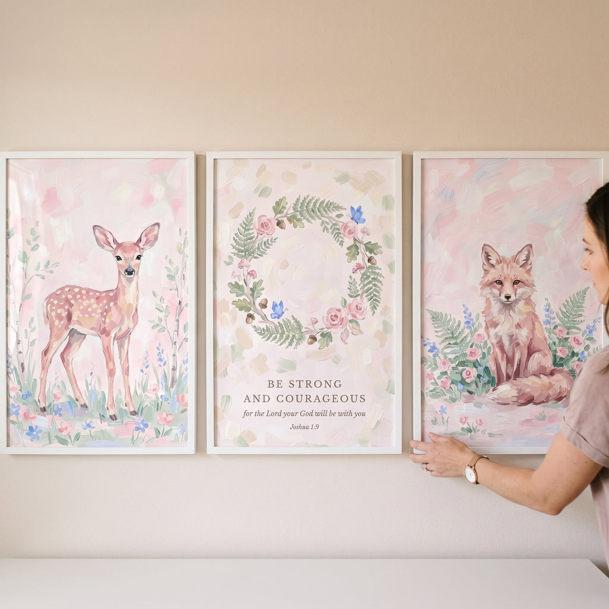 A person hangs a grandmillennial blush woodland nature wall art set of 3 with watercolor deer, fox, and wreath designs.
