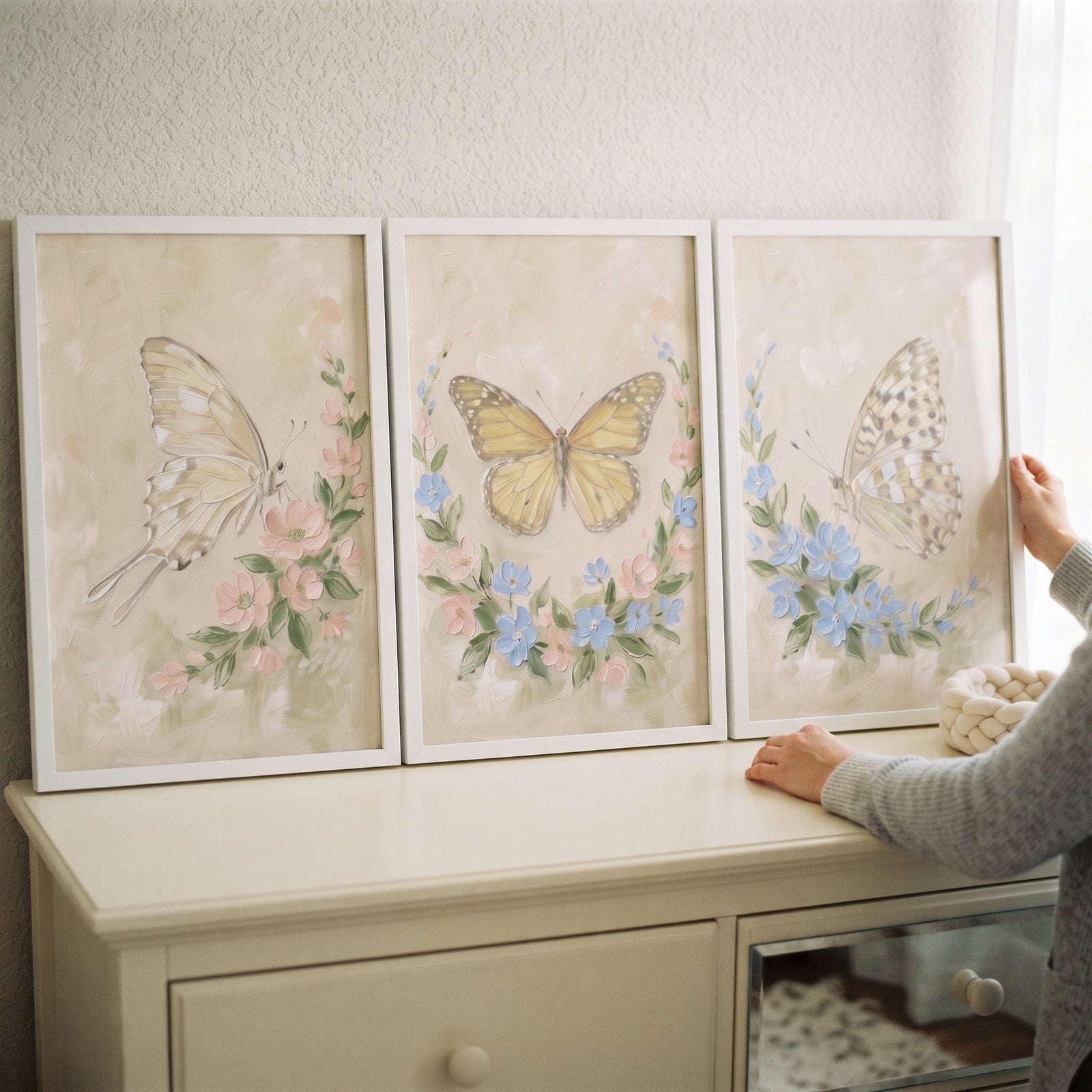 A hand touches a grandmillennial floral garden butterfly wall art set of 3 on a white dresser.