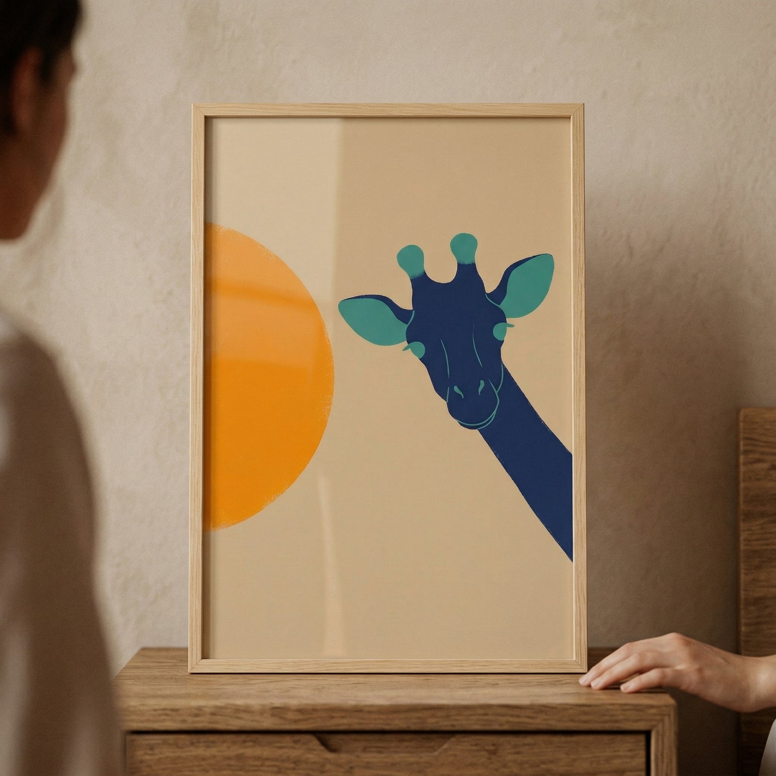 Framed minimalist giraffe illustration with a blue giraffe head peeking in from the side beside a large orange sun. The artwork rests on a wooden dresser with a hand adjusting the frame, adding a styled home decor feel.