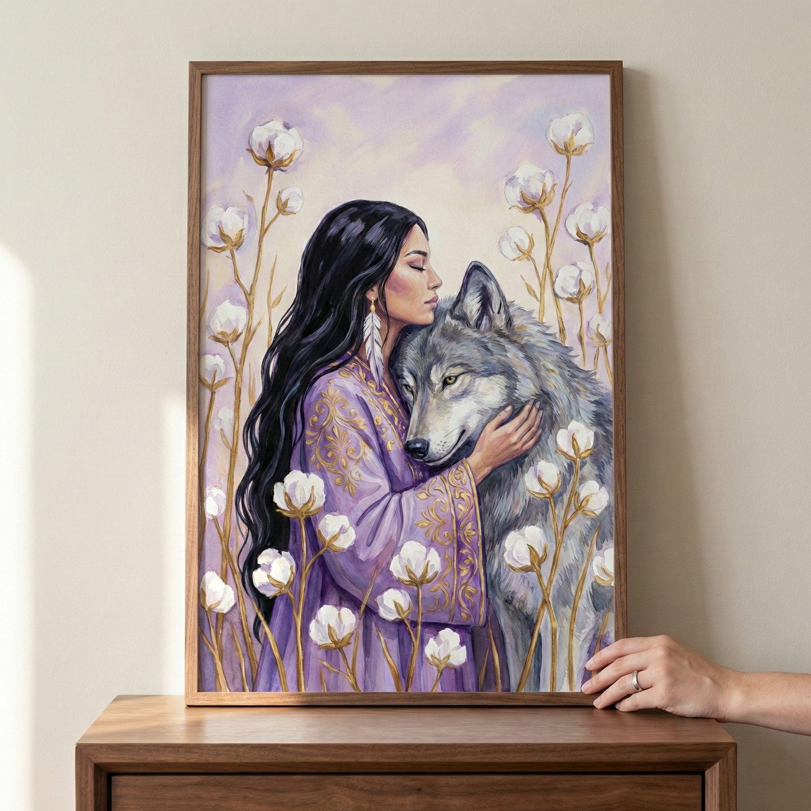 A framed artwork showing a woman with long black hair and closed eyes in a tender embrace with a large grey wolf. The woman wears a lavender robe with intricate gold patterns and a feather earring. They are surrounded by tall cotton plants with white bolls under a pale purple sky. The frame is being held by a hand at the side, resting on a dark wood cabinet against a light-colored wall.