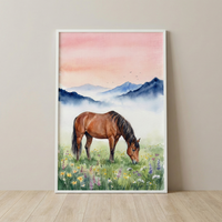 A framed piece of watercolor artwork leaning against a beige wall on a light wood floor. The illustration features a brown horse with a black mane and tail grazing in a meadow filled with colorful wildflowers in shades of purple, yellow, and white. Behind the horse, soft mist clings to the base of layered blue mountains. The sky is a gentle wash of pink and orange, with several small bird silhouettes flying in the distance.