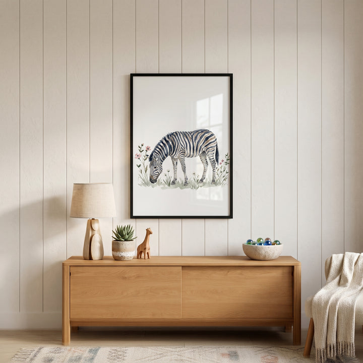 Grazing meadow zebra wall art hangs above a wooden sideboard with a lamp and small decor on a white paneled wall.