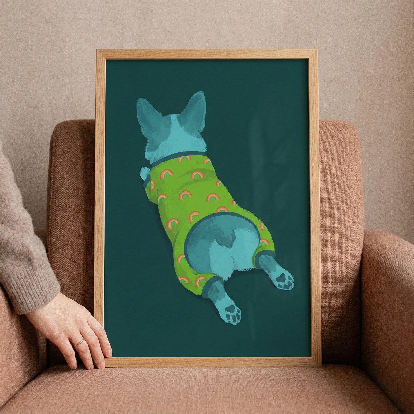 A framed illustration of a blue dog seen from behind, lying flat on its belly with its legs spread out. The dog wears a lime green onesie decorated with orange rainbow motifs. The artwork features a deep teal background and is set within a light-colored wooden frame. 