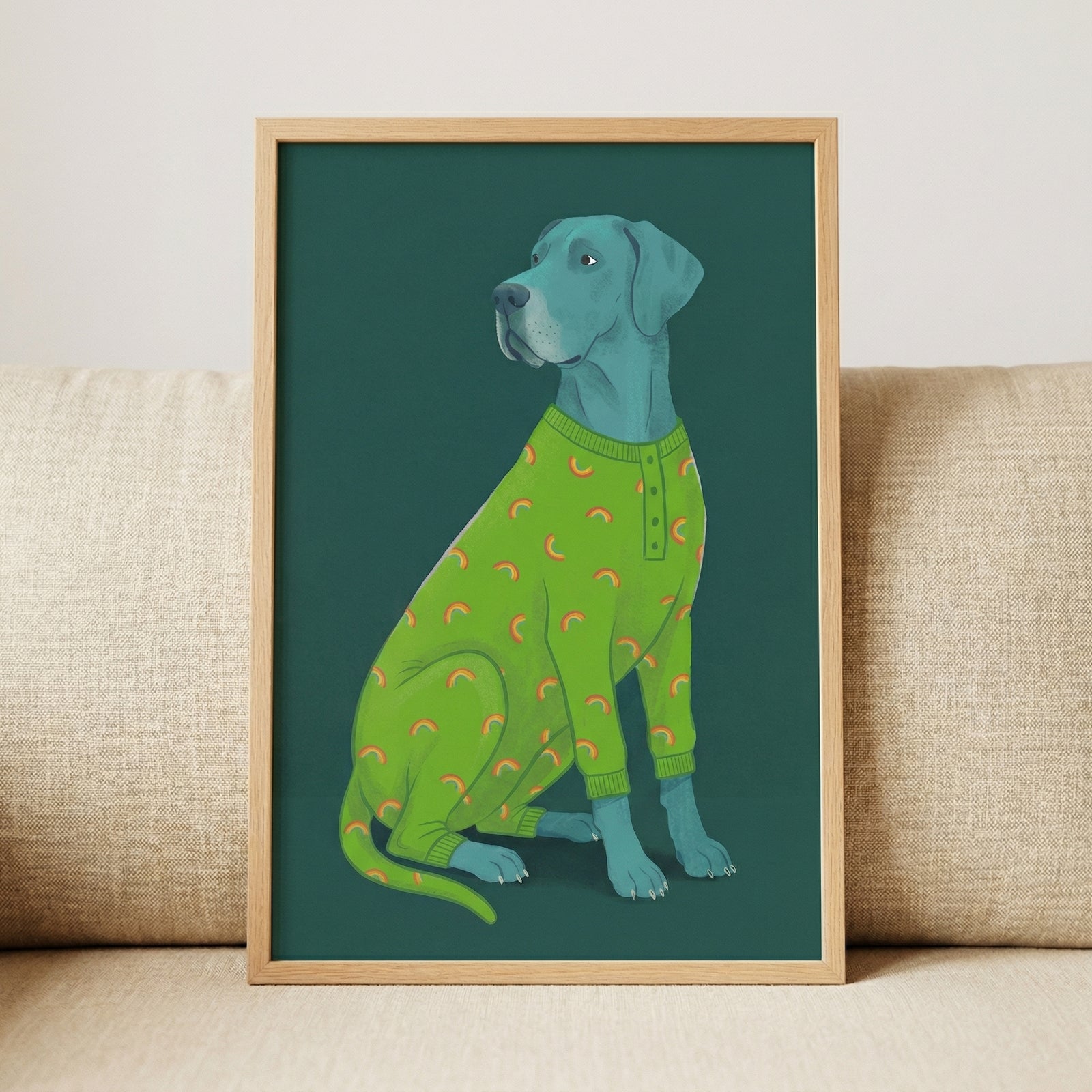 A digital illustration of a large, blue-gray dog sitting and looking over its shoulder. The dog is dressed in bright green pajamas featuring a recurring pattern of small orange and yellow rainbows. The illustration has a clean, graphic aesthetic and is set against a solid dark teal background. The framed artwork, in a light-toned wooden frame, is resting on a neutral-colored, textured couch.