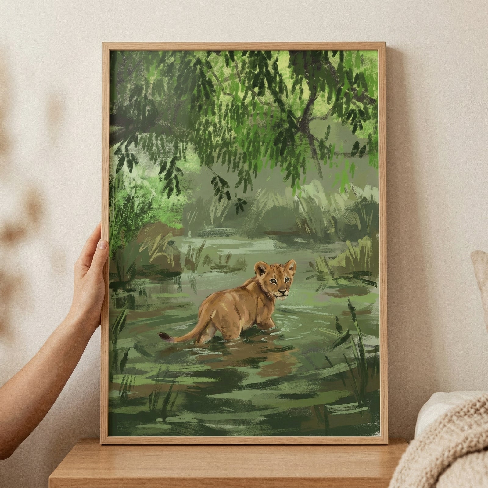 A digital illustration shows a young lion cub wading through a shallow river in a lush jungle. The cub looks back over its shoulder, its golden fur contrasting with the deep greens of the surrounding foliage. Overhanging leafy branches create a canopy above. The artwork uses broad, expressive brushstrokes to depict the scene, with a focus on light and color. The framed print is held by a hand and placed on a wooden surface against a neutral wall.