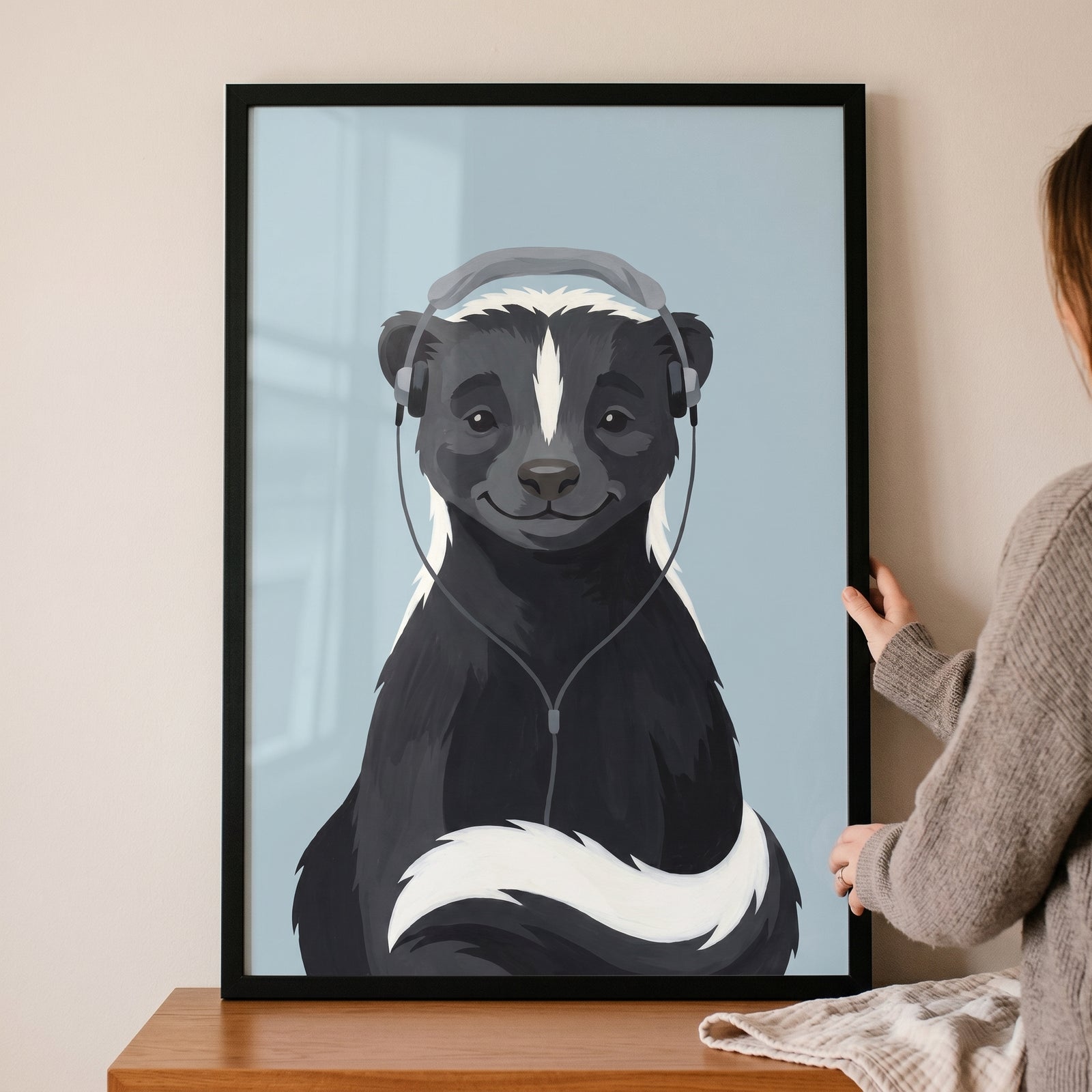 Framed grey headphone listening skunk wall art held by a person, showing a cute skunk against a light blue backdrop.