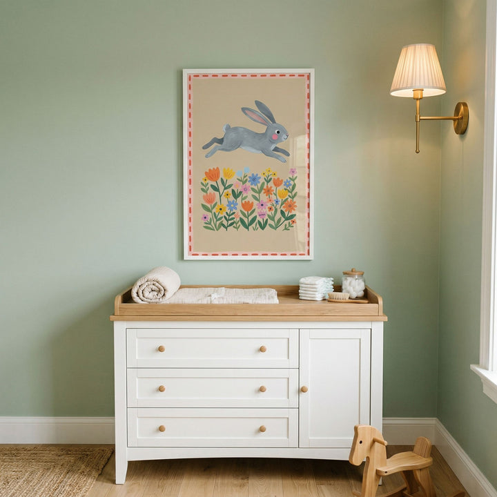 Grey meadow jumping bunny wall art above a white changing table in a sage nursery with a gold lamp and rocking horse.