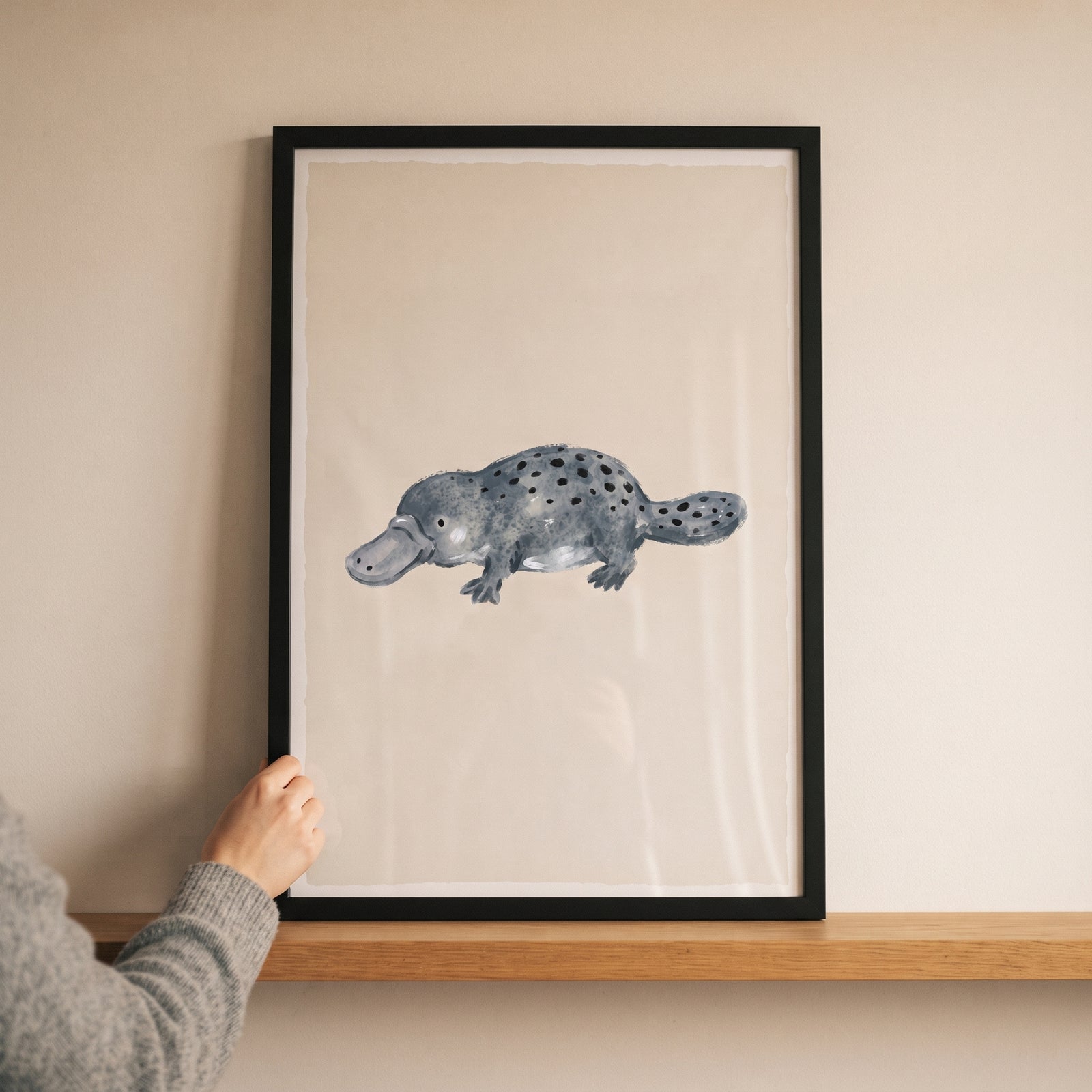 Hand holding a black frame featuring grey spotted platypus wall art on a wooden shelf against a beige wall.