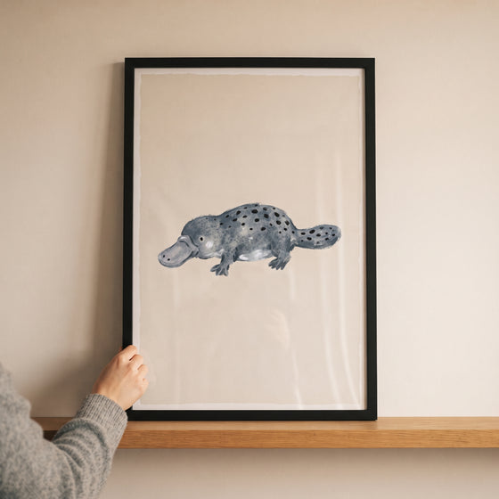 Hand holding a black frame featuring grey spotted platypus wall art on a wooden shelf against a beige wall.