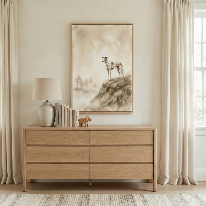 This minimalist interior scene centers on a large, vertically oriented framed artwork hanging on a warm beige wall. The artwork features a slender, spotted sighthound standing atop a craggy cliff, looking out over a hazy, atmospheric landscape filled with soft mist and a few simplified evergreen tree shapes. The piece is rendered in a muted palette of earth tones, including tan, sepia, and cream.

