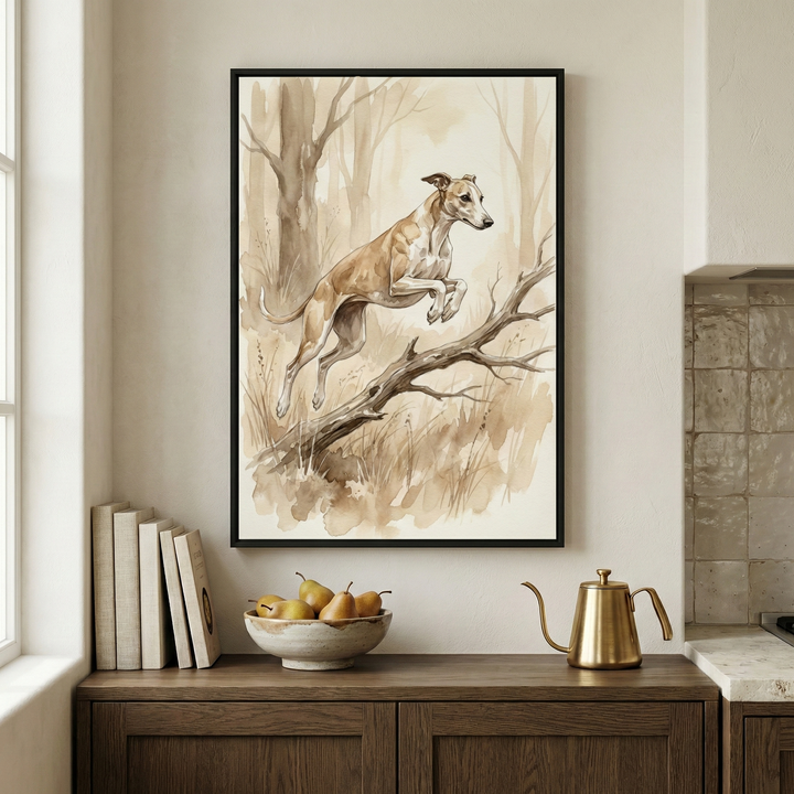 A vertical art piece in a slim black frame hangs on a neutral wall. The artwork is a watercolor-style illustration of a slender tan-and-white dog leaping over a fallen tree limb in a minimalist forest setting, rendered in muted sepia and brown tones. Below the frame, a dark wood cabinet holds a stack of plain books, a ceramic bowl filled with yellow pears, and a golden metallic kettle with a long, curved spout. A tiled wall and kitchen countertop are visible to the right.