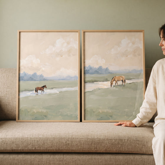 Hazy creek meadow horse wall art set of 2 framed prints displayed on a textured sofa next to a person in white.