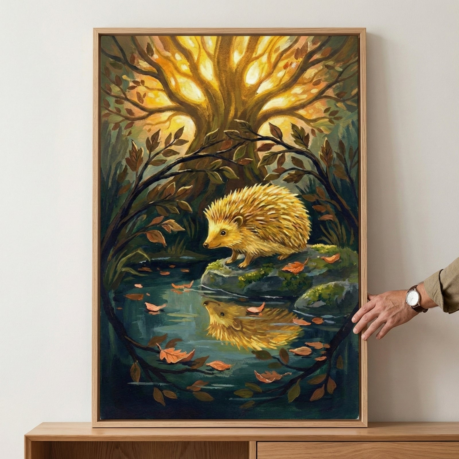 A framed vertical illustration depicts a golden-furred hedgehog standing on a mossy stone by a dark, reflective pool. Its reflection is visible in the water, which is dotted with orange and brown fallen leaves. In the background, a massive tree with intricate branches glows with a warm, golden light. A circular frame of leafy branches surrounds the central scene. To the right, a person's hand with a wrist watch holds the side of the light-colored wooden frame. The overall style is detailed and magical, with