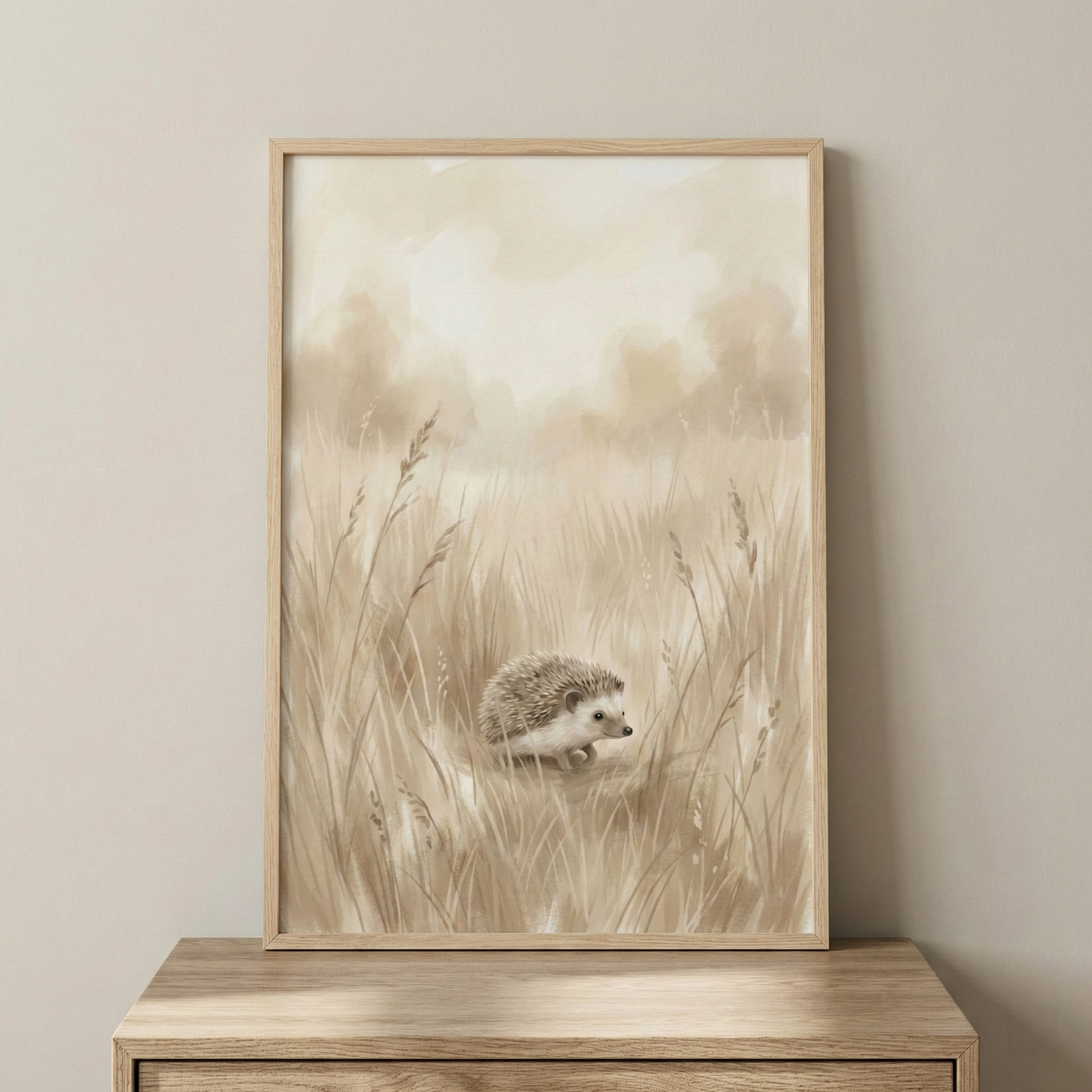 A framed piece of wall art features a gentle illustration of a small hedgehog in a field of tall grass. The style is soft and minimalist, using a muted color palette of cream, beige, and light brown tones. The hedgehog is depicted in a side profile, walking through the wispy grass which reaches up towards a hazy, light-colored background. The image is presented in a light wood frame, resting on a wooden tabletop against a neutral-colored wall. The overall aesthetic is calm, delicate, and earthy.