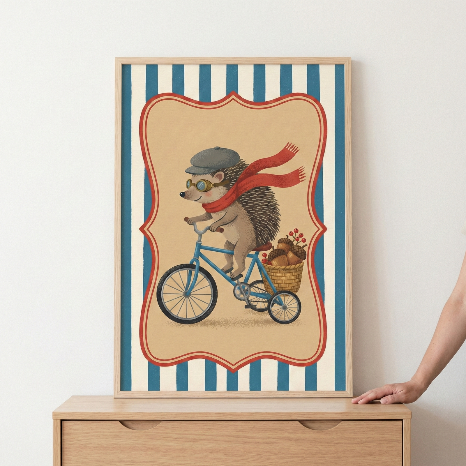 A vertical framed illustration sits on a light wood dresser against a plain white wall. The artwork depicts a whimsical hedgehog riding a blue bicycle. The animal is outfitted with a grey flat cap, round goggles, and a long red scarf trailing behind it in the wind. A small wicker basket attached to the back of the bike is filled with acorns and red berries. The central scene is enclosed in a decorative red border, set against a background of vertical blue and white stripes. 