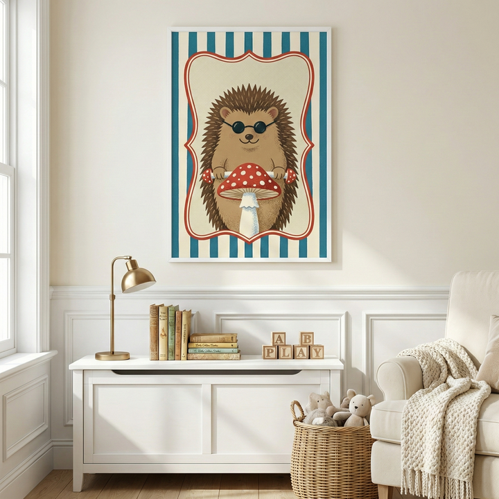 A framed piece of art hangs on a cream-colored wall above a white wooden storage chest in a sunlit room. The illustration features a brown hedgehog wearing round black sunglasses, sitting behind a large red mushroom with white spots. The hedgehog is centered within a decorative red scalloped border against a background of blue and white vertical stripes. On top of the chest below the art is a gold lamp, a row of colorful books, and wooden blocks that spell "PLAY". To the right of the chest, a woven basket h