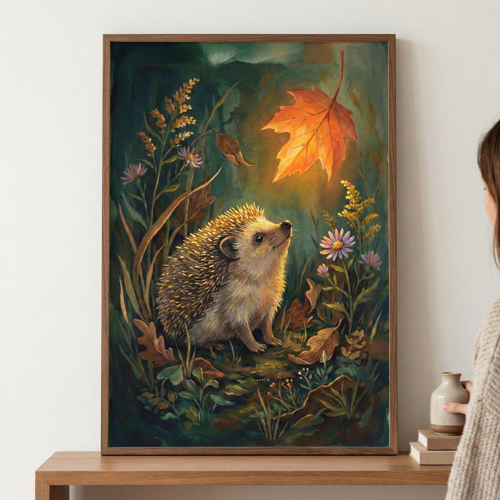 A framed illustration featuring a small hedgehog gazing up at a large, luminous orange autumn leaf. The scene is filled with dark green foliage, delicate purple flowers, and dried leaves. The glowing leaf provides a warm, central light source in the dark woodland setting. The framed artwork is displayed on a light-colored wall above a wooden table.