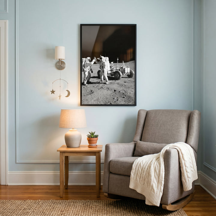 A grey rocker sits next to a wooden side table beneath framed high five moon astronaut wall art on a blue wall.