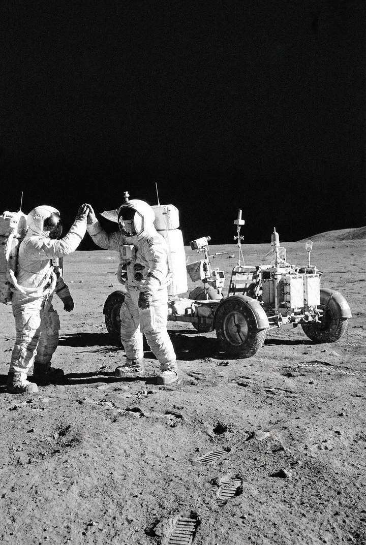 Two astronauts in full space suits stand on the moon raising their hands toward each other beside a lunar rover, with footprints visible across the dusty surface. The dark sky contrasts sharply with the bright textured ground, emphasizing human presence in space exploration.

