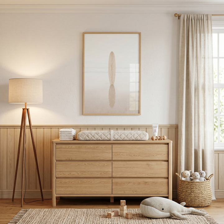 High key minimalist surfboard wall art above a wood dresser in a nursery with a tripod lamp and plush whale toy.