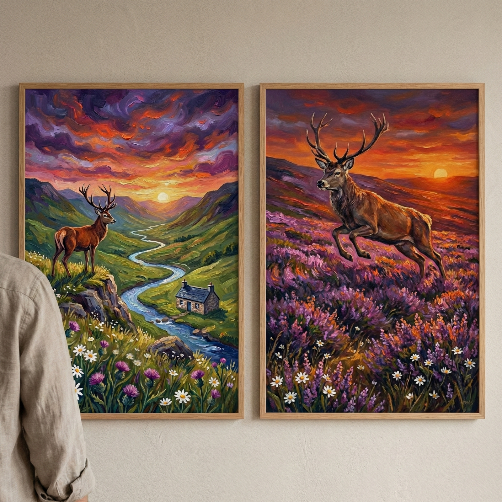 A pair of framed artworks hang side-by-side on a neutral-toned wall. The artwork on the left shows a stag standing on a rocky ledge overlooking a deep green valley. A winding blue river flows past a small stone cottage, and hills rise in the distance under a vibrant sunset with purple and orange clouds. The artwork on the right features a stag mid-leap across a field of purple heather and white flowers. 