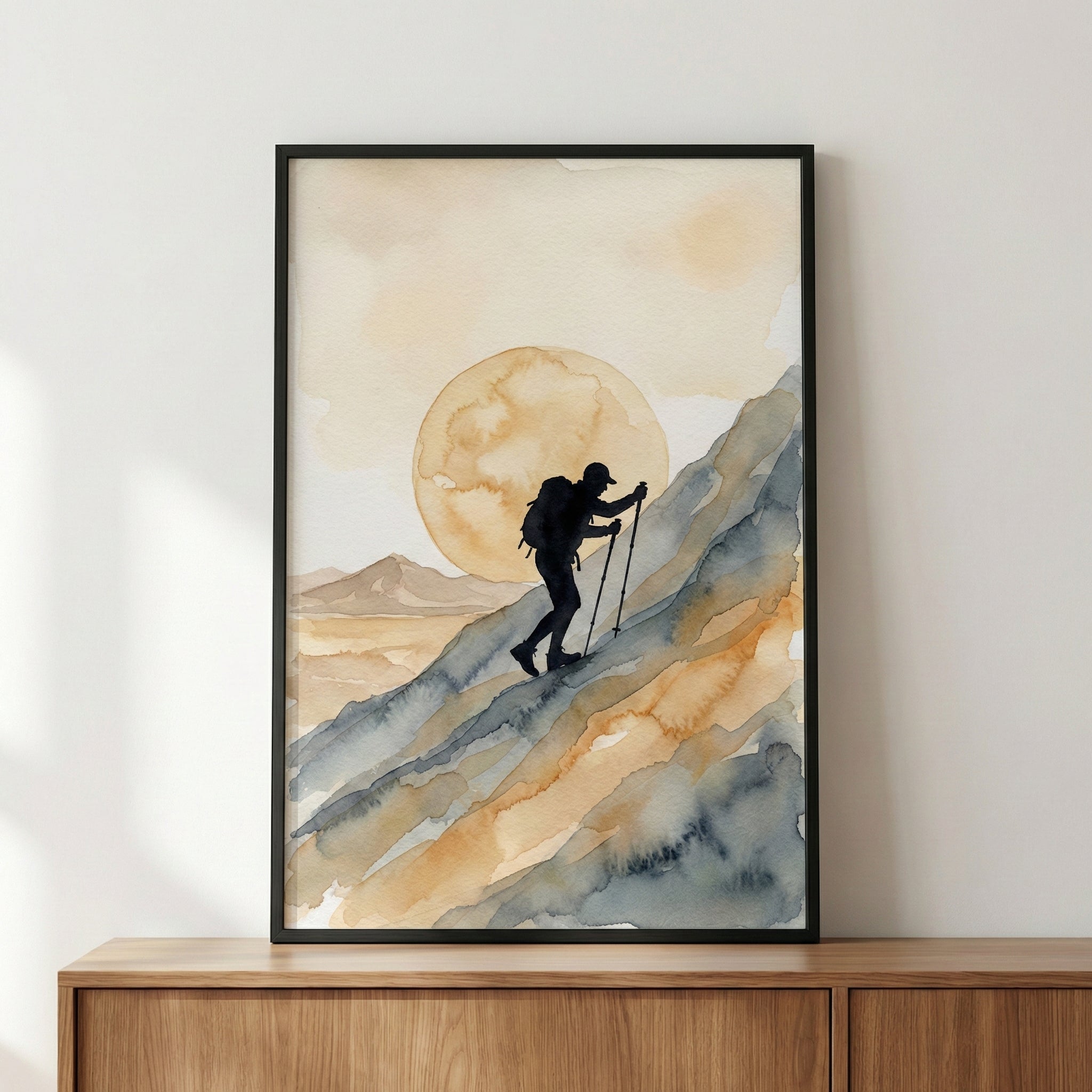Framed watercolor painting of a hiker climbing a steep mountain slope with trekking poles, silhouetted against a large, golden sun in the background. The artwork features soft, earthy tones of blue, brown, and beige, blending to create a serene and adventurous landscape.
