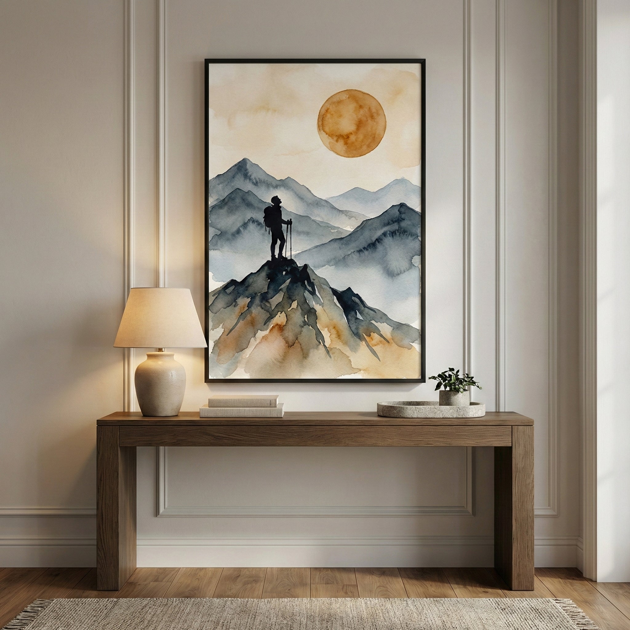 Modern living room with a large framed watercolor painting of a lone hiker standing on a cliff, facing distant mountain peaks and a glowing golden sun. The room features a dark wood sideboard, pampas grass in a tall vase, a stack of design books, and a brass table lamp, creating a warm and earthy aesthetic.
