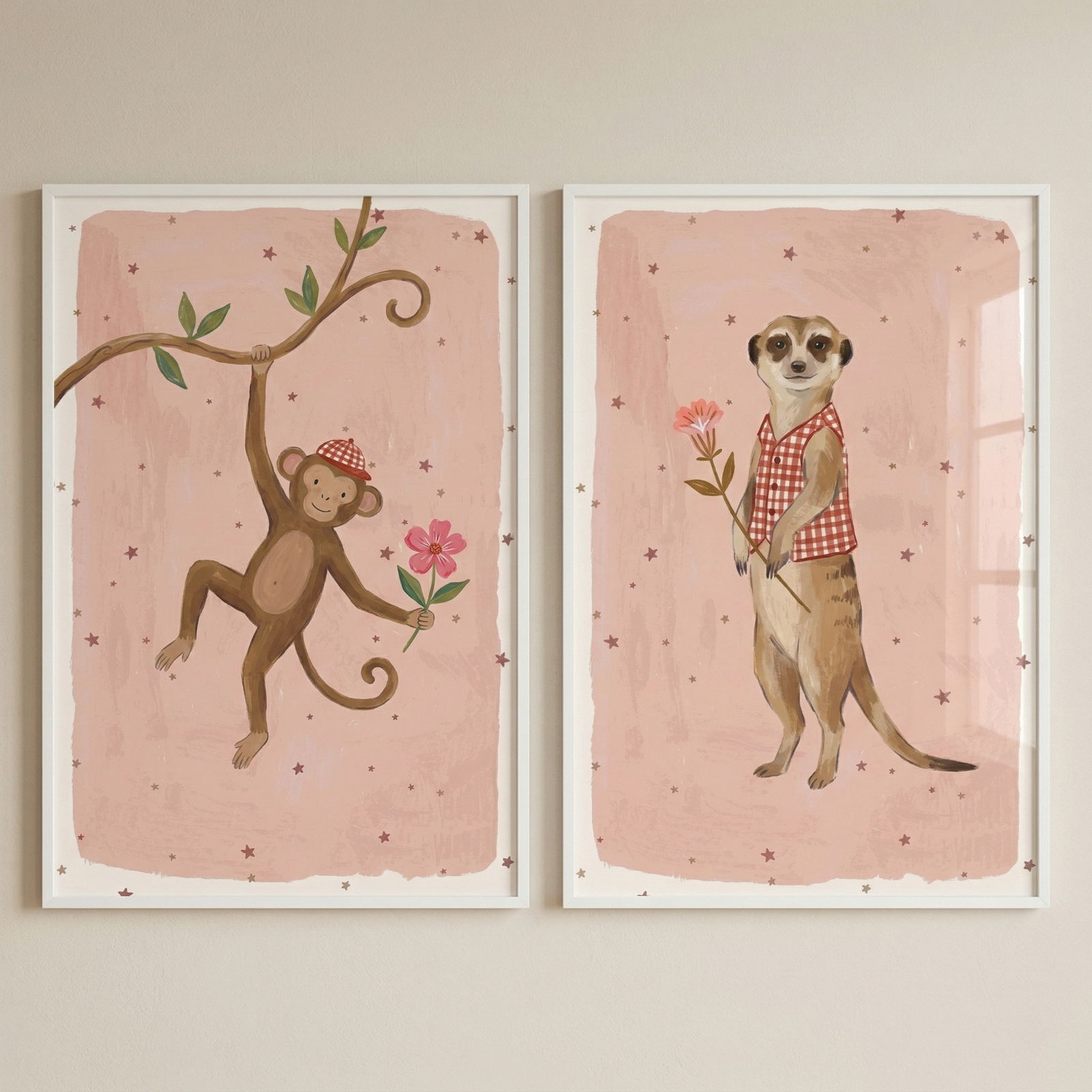 Two framed illustrations hang side-by-side. The left artwork shows a cartoon monkey swinging from a branch, wearing a red checkered hat and holding a pink flower. The right artwork features a meerkat standing upright in a matching checkered vest, also holding a pink flower. Both have a soft pink, star-patterned background and are set within slim white frames. The whimsical style is ideal for a child's bedroom or nursery decor.