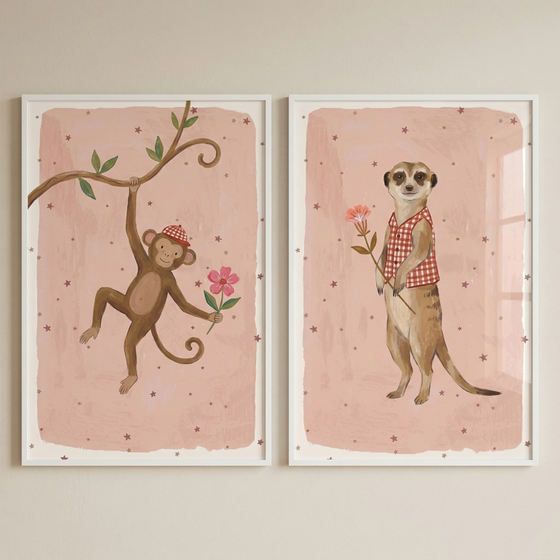 Two framed illustrations hang side-by-side. The left artwork shows a cartoon monkey swinging from a branch, wearing a red checkered hat and holding a pink flower. The right artwork features a meerkat standing upright in a matching checkered vest, also holding a pink flower. Both have a soft pink, star-patterned background and are set within slim white frames. The whimsical style is ideal for a child's bedroom or nursery decor.