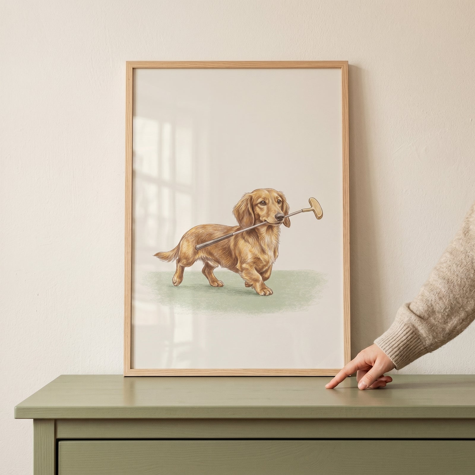 Illustration of long-haired dog, holding golf putter dachshund dog wall art, in a wooden frame on a green dresser.