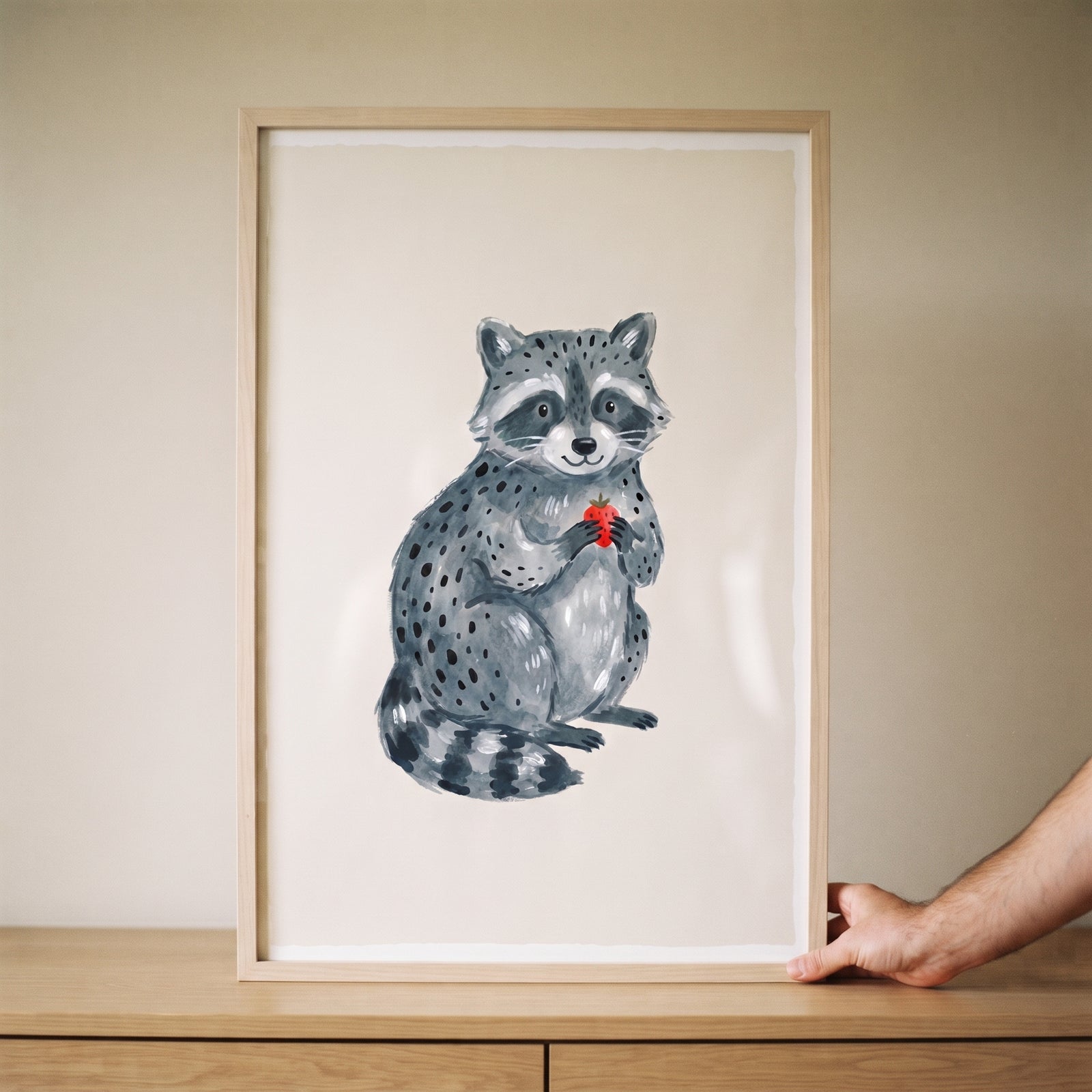 Framed holding strawberry grey raccoon wall art on a wooden surface, supported by a hand against a neutral background.