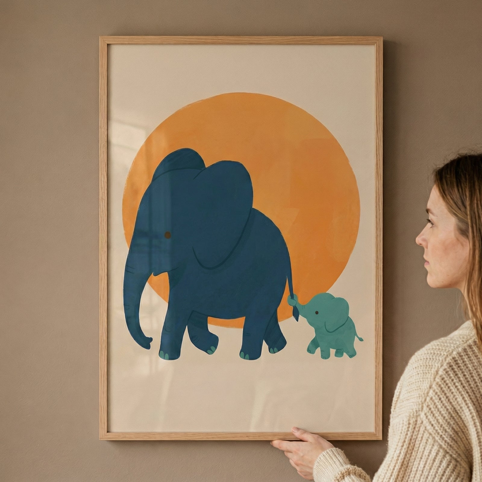A framed illustration shows a large blue elephant and a small baby elephant walking together in front of a warm orange circle. A person holds the frame against the wall, highlighting it as a playful and gentle nursery art piece.