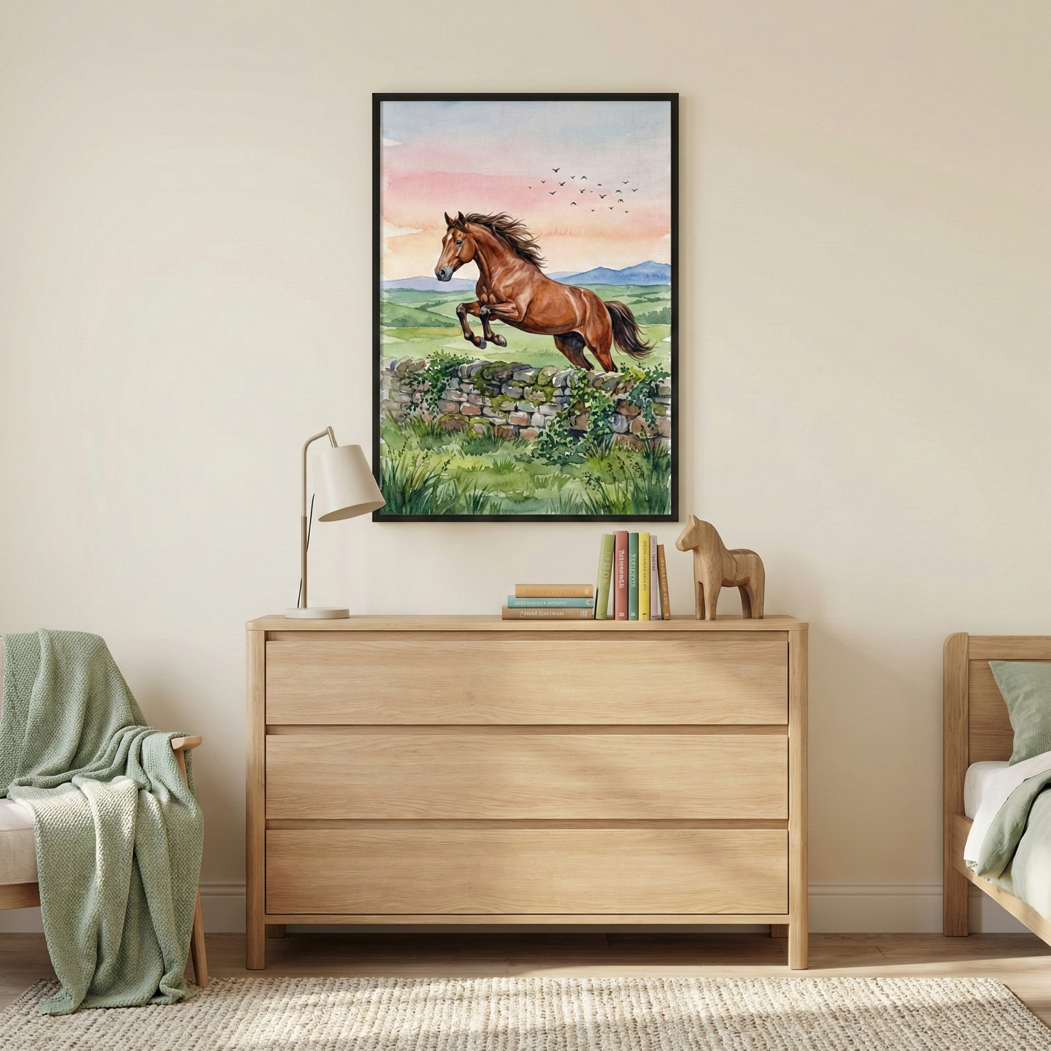 A framed piece of art hangs on a neutral-toned wall above a light-colored wooden three-drawer chest. The artwork depicts a brown horse in mid-air as it jumps over a low stone wall. The landscape features lush green fields and rolling hills under a soft sunset sky of pink and orange hues, with a small flock of birds flying in the distance. 


