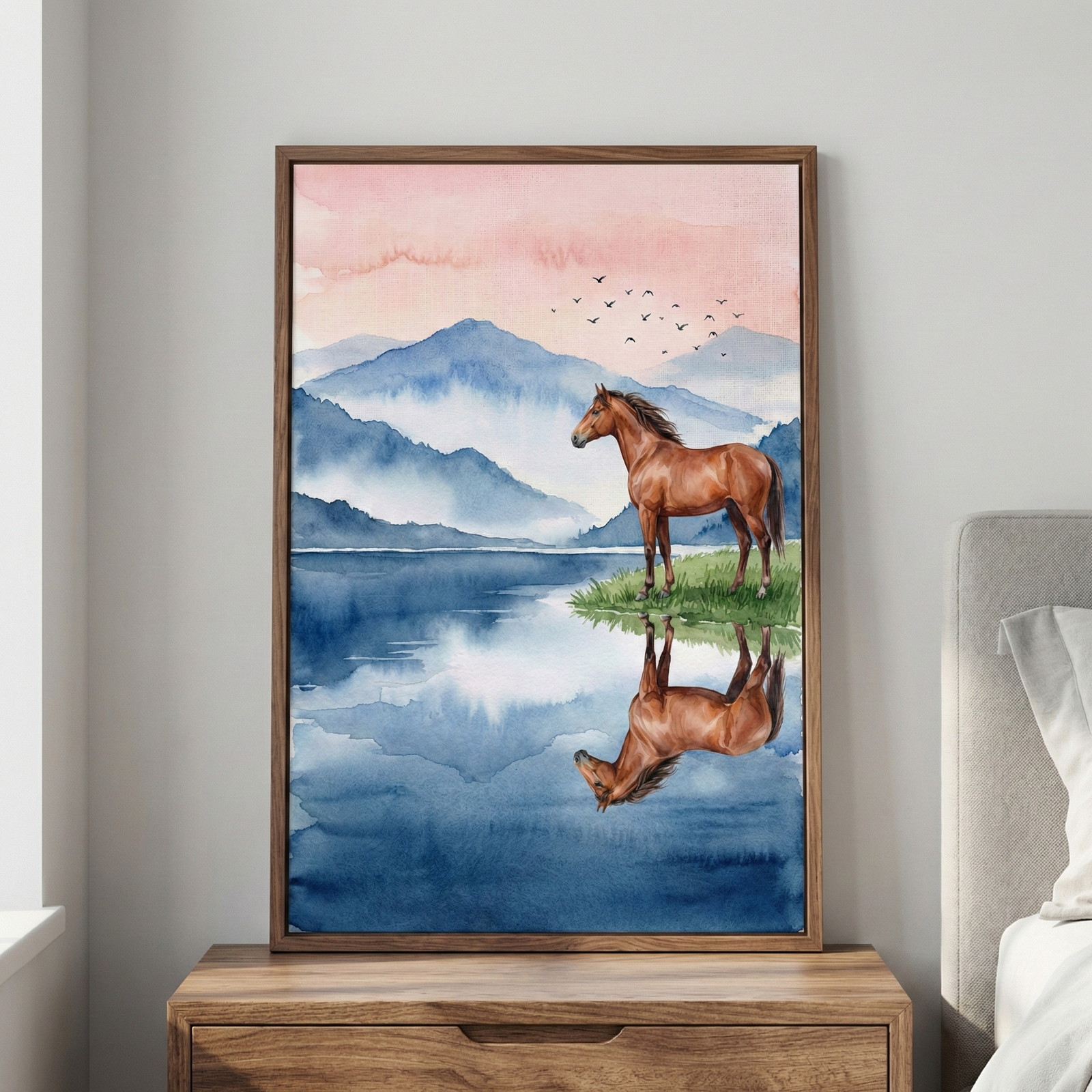 A framed watercolor-style artwork hangs on a light-colored wall above a wooden chest of drawers. The vertical piece depicts a brown horse standing on a grassy bank beside a calm body of water. A clear reflection of the horse is visible in the dark blue water below. The background features several layers of misty blue mountains under a soft pink and orange sky. A small flock of birds is seen flying in the distance. The artwork is held in a minimalist brown wooden frame.