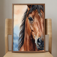 A framed watercolor illustration of a brown horse's head sits on a light wooden chair against a neutral wall. The close-up portrait shows the horse's expressive dark eye, detailed muzzle, and flowing dark mane. The artwork's background consists of soft, blended washes of pale orange and blue. The piece is encased in a simple, thin wooden frame that matches the natural wood of the chair it's propped on.