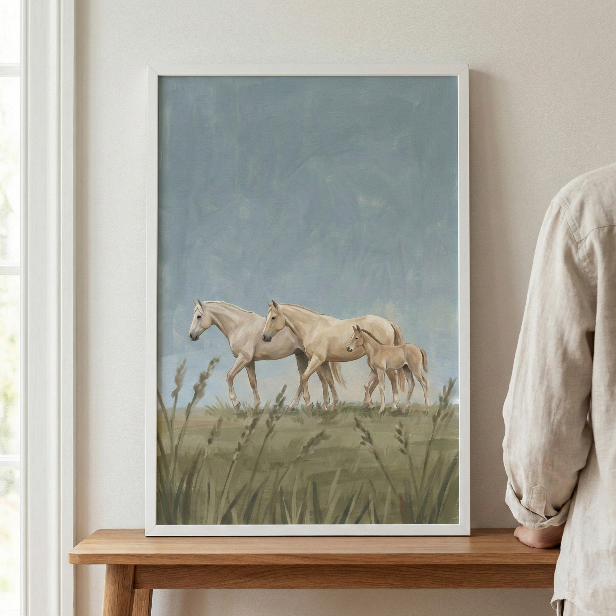 A framed vertical illustration shows three light cream-colored horses—two adults and a foal—walking through a field of tall grass. The artwork features a soft, muted color palette with a cloudy, textured sky in shades of blue and grey. The piece is in a minimalist white frame, resting on a light wood surface against a neutral wall. A person in a linen shirt is partially visible on the right, showing the artwork in a home setting.