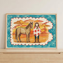Framed art print of a girl and horse against a vibrant sunset, propped on a light wood shelf with neutral wall tones. The colorful and rustic wall art brings a playful, storybook feel to modern interiors.