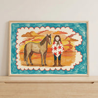 Framed art print of a girl and horse against a vibrant sunset, propped on a light wood shelf with neutral wall tones. The colorful and rustic wall art brings a playful, storybook feel to modern interiors.
