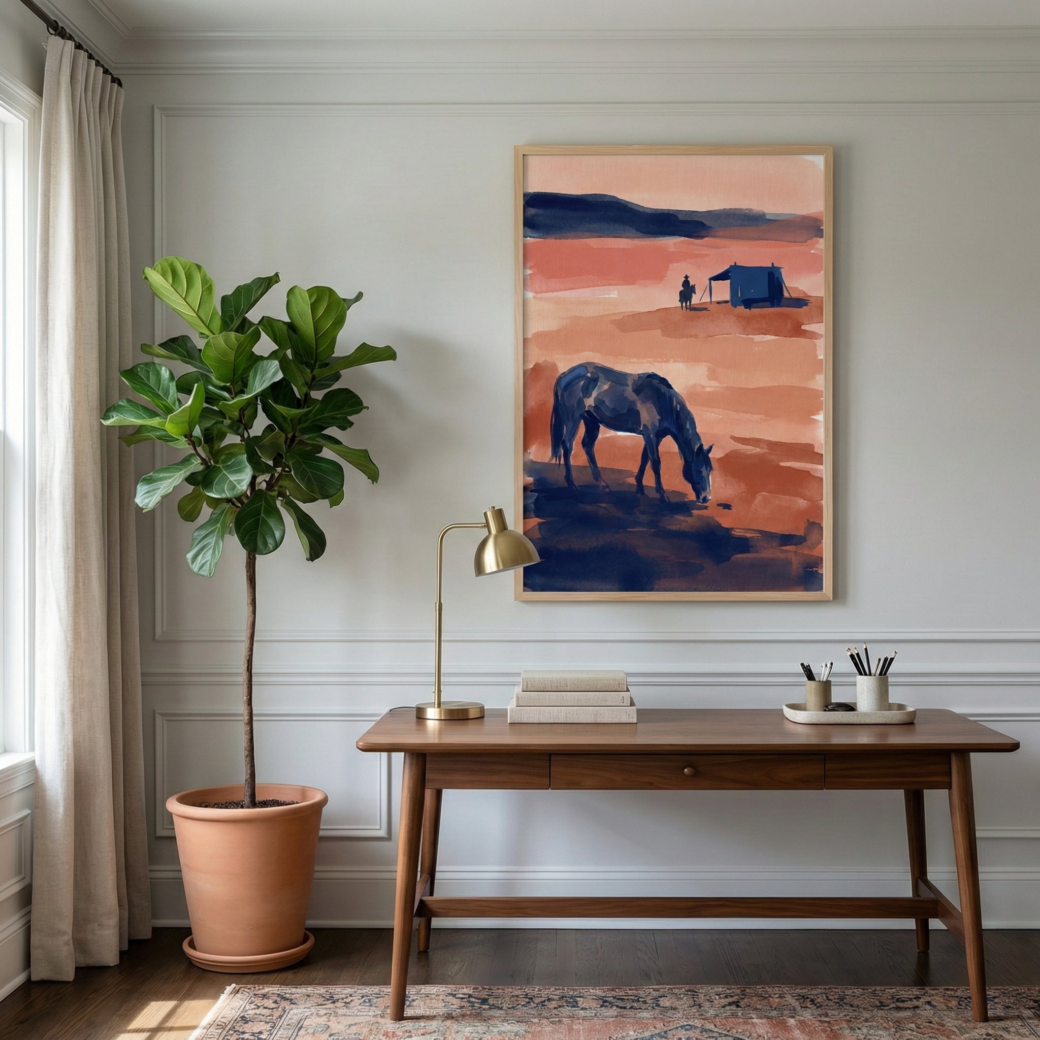 Framed desert scene artwork featuring a grazing horse and a distant cowboy displayed above a mid-century modern desk in a bright, stylish home office with a fiddle-leaf fig plant.