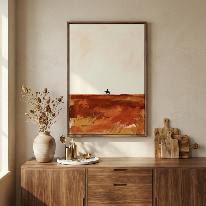 Framed western desert artwork featuring a silhouette of a cowboy on horseback, displayed above a wooden sideboard with neutral decor and warm lighting in a modern rustic interior.