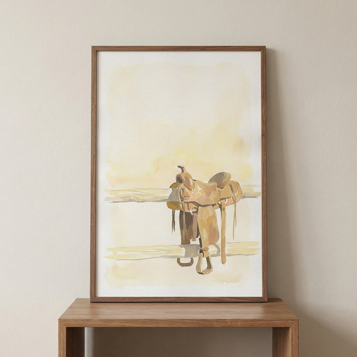 Framed watercolor-style artwork of a western horse saddle resting on a wooden rail, rendered in soft beige and brown tones. The minimalist composition emphasizes the iconic silhouette of cowboy gear against a light, sunlit background.
