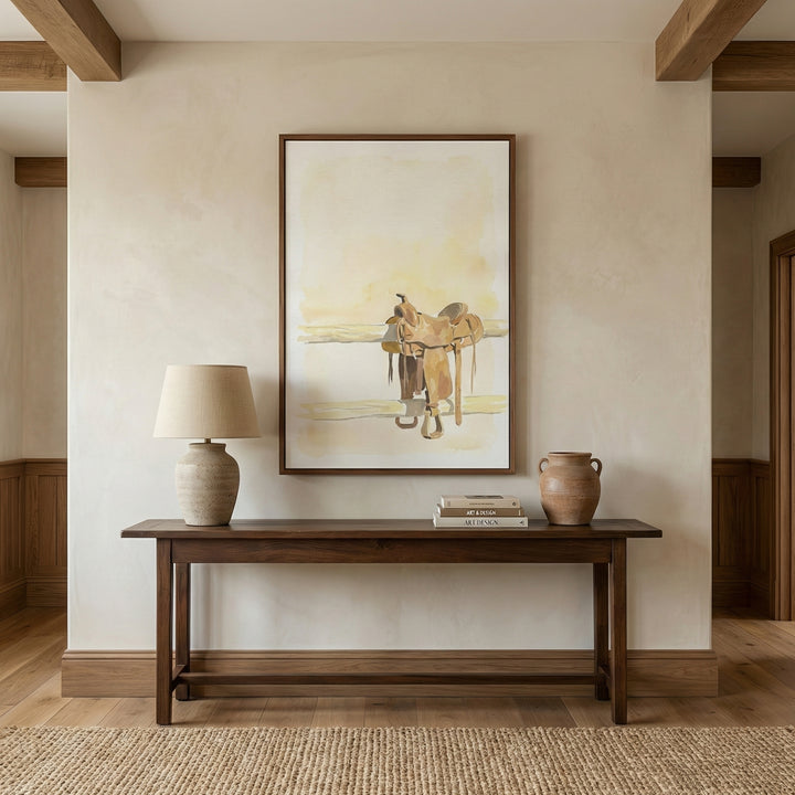 Large western-themed art print featuring a watercolor horse saddle on a fence, displayed in a rustic hallway above a wooden console table. The painting evokes cowboy culture with warm, neutral hues and a quiet, open-range feel.
