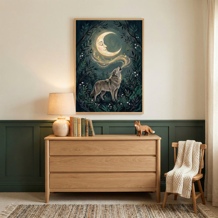 A cozy interior scene featuring a large framed piece of art on a wall above a light wood dresser. The art depicts a grey wolf in a forest howling towards a golden crescent moon with a serene face, with a trail of stardust between them. On the dresser sits a round table lamp with a beige shade, several books, and a small wooden fox figurine. To the right, a wooden chair has a thick cream-colored blanket draped over its back. The wall is cream on the top half and dark forest green on the bottom half. A patter