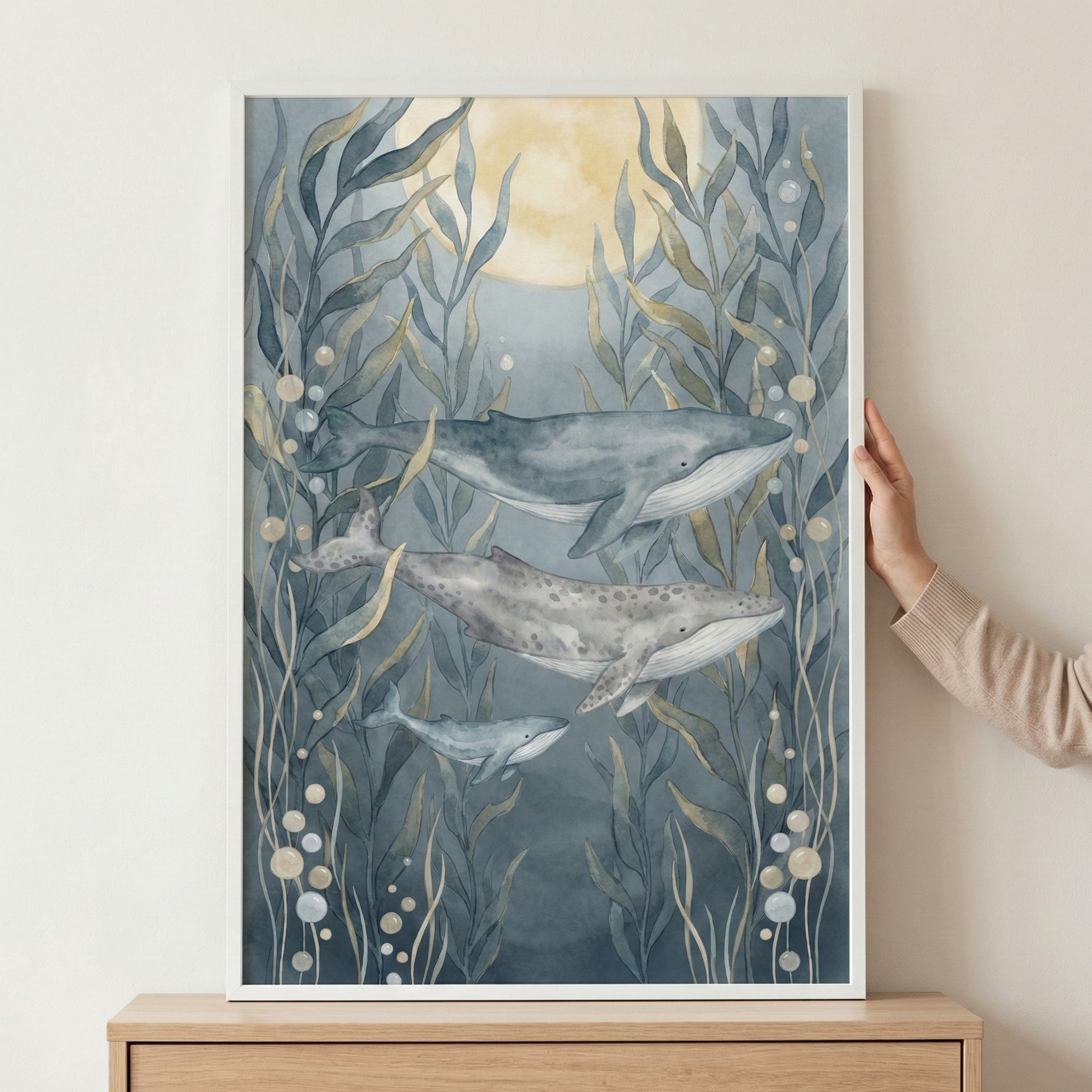 A framed watercolor-style illustration depicts three whales swimming through a serene underwater forest. Tall, flowing aquatic plants in shades of blue and muted green frame the scene, accented by small floating bubbles. In the upper background, a large, soft yellow moon glows through the deep blue water. The artwork is housed in a white frame, which is held by a person's hand as it rests on a light wooden surface against a neutral wall.