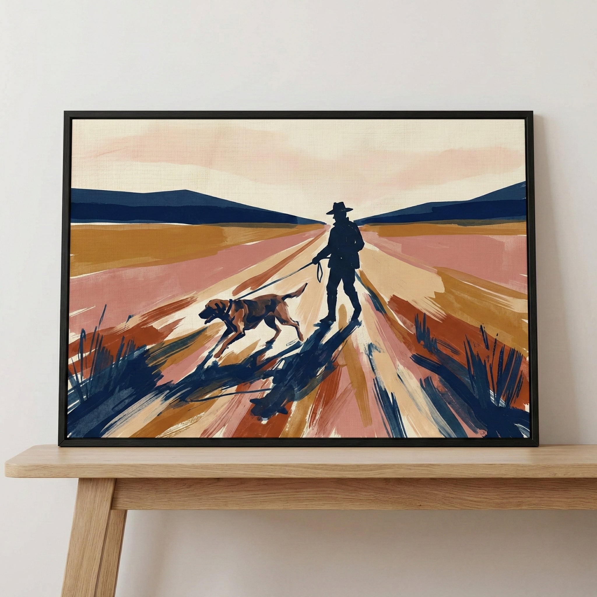 A framed illustration of a man in a hat walking a dog on a leash along a wide, perspective-driven path. The landscape is rendered in expressive, broad brushstrokes with a color palette of warm ochre, pink, and orange, contrasted by deep blue mountains in the distance. Bold, dark strokes create texture and shadows across the scene. The artwork is shown in a black frame, resting on a light wood table against a neutral wall.