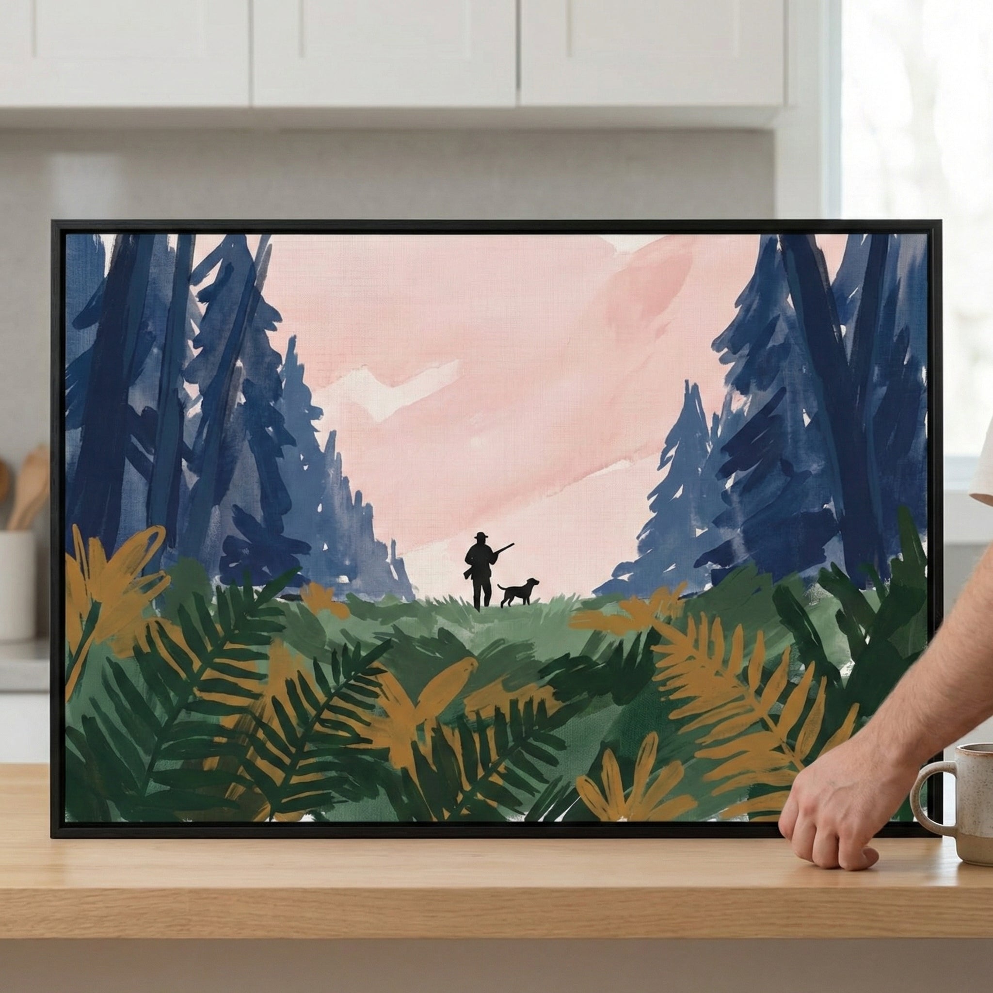 A framed illustration displayed on a light wood surface, depicting a stylized outdoor scene. In the center distance, the dark silhouettes of a person holding a long-barreled item and a dog are visible in a clearing. The scene is bordered by tall, dark blue trees against a pale pink sky. The foreground is filled with large, layered fronds in shades of deep green and golden yellow. A person's hand and a mug are visible to the right of the frame, set against a background of blurred white kitchen cabinets.