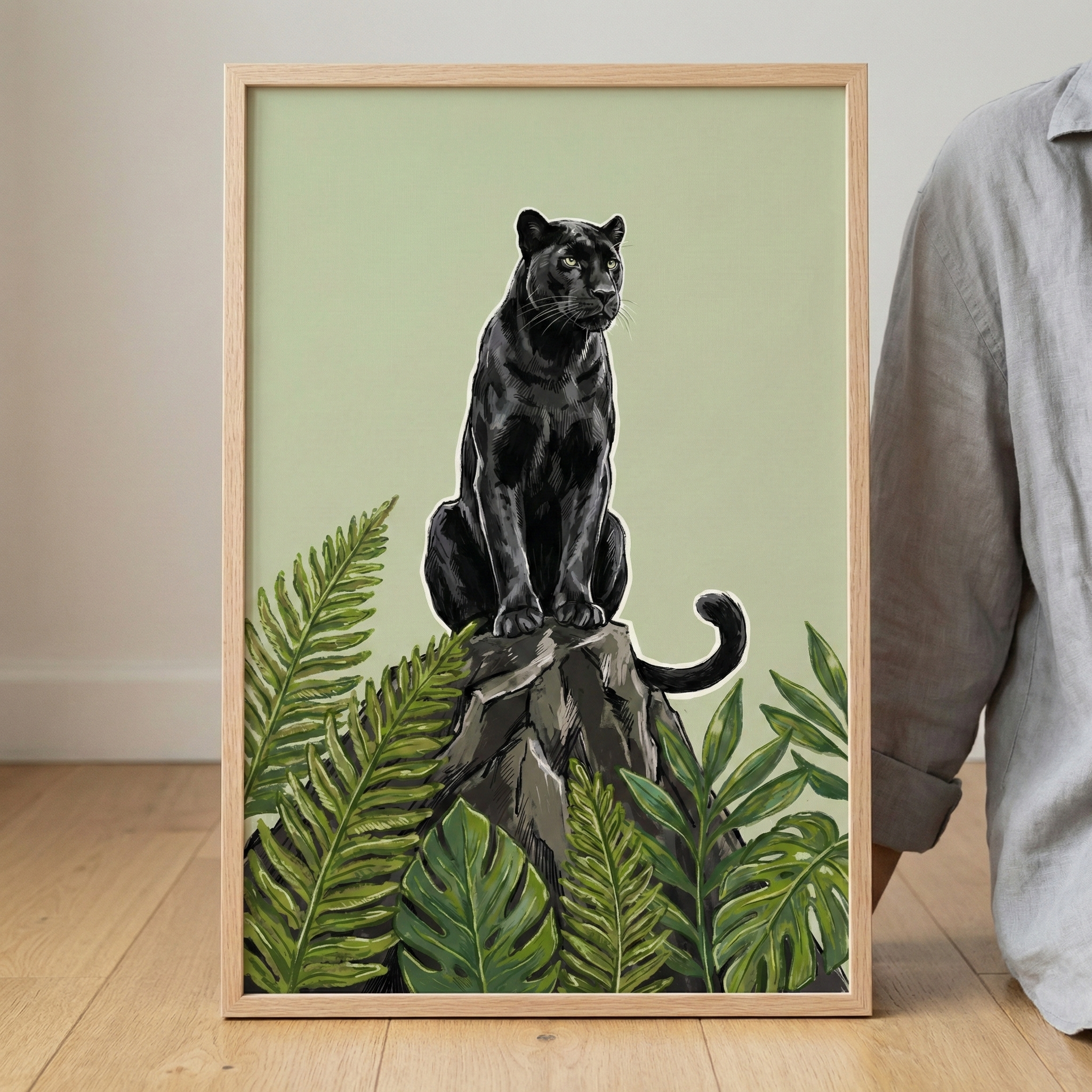 A framed art print features a sleek black big cat sitting upright on a textured grey rock. The animal is surrounded by lush green tropical foliage, including large ferns and broad leaves at the base. The graphic illustration is set against a solid, pale olive-green background. The print is held in a light wood frame and leans on a light-colored wooden floor against a plain white wall, with a person in a grey shirt partially visible on the right.