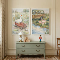 Two framed illustrations hang on a cream-colored wall above a light green two-drawer dresser. The art on the left features a large white goose sitting in a small red pedal car, with a tiny green frog at the wheel holding a small white flag. They are depicted in a serene natural setting with tall grass and wildflowers. 
