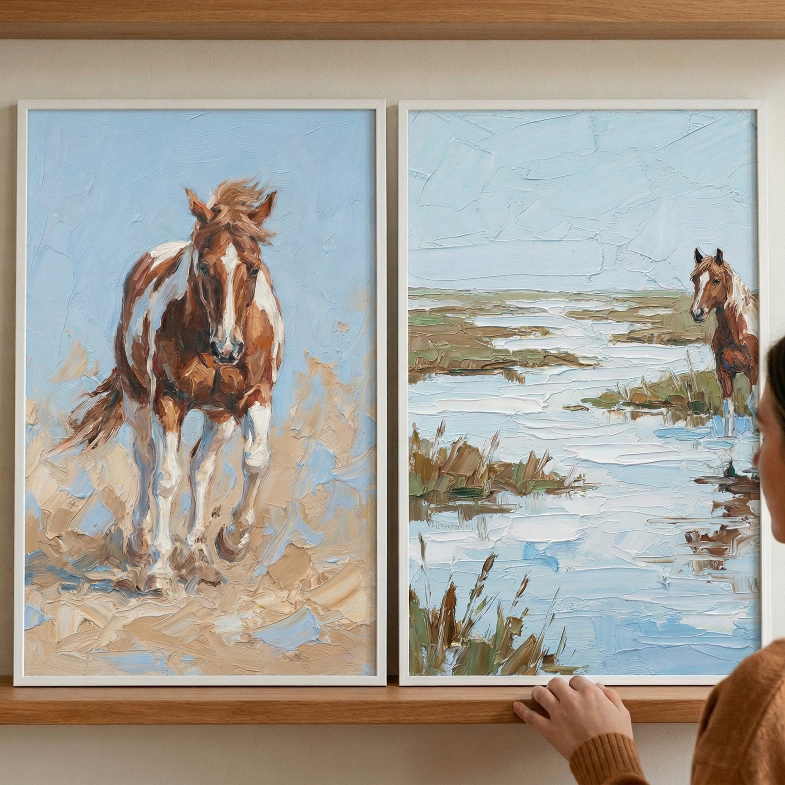 Two framed paintings rest on a wooden shelf, one showing a brown and white horse walking forward with textured brushstrokes and the other showing a horse standing in a marshy landscape with water and grasses. A person’s hand reaches toward the frame, suggesting the artwork is being arranged or styled on the wall.